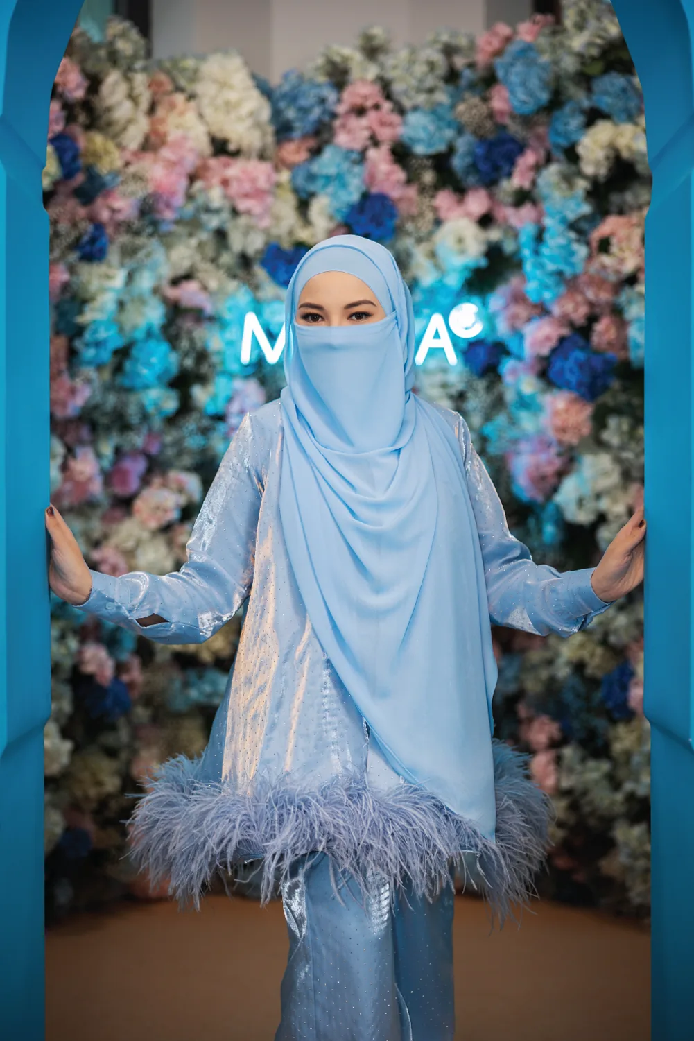 Neelofa in a powder-blue look framed by a blue archway and florals.