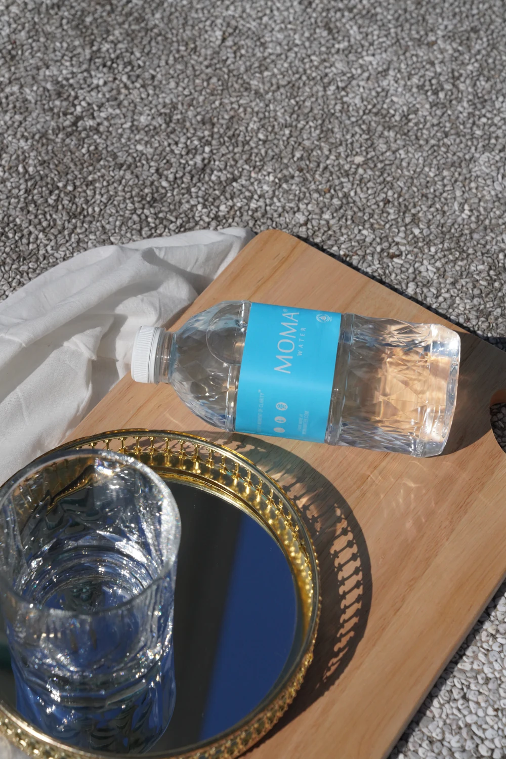 A MOMAwater bottle resting on a wooden service board, a glass on a gilded tray beside it.