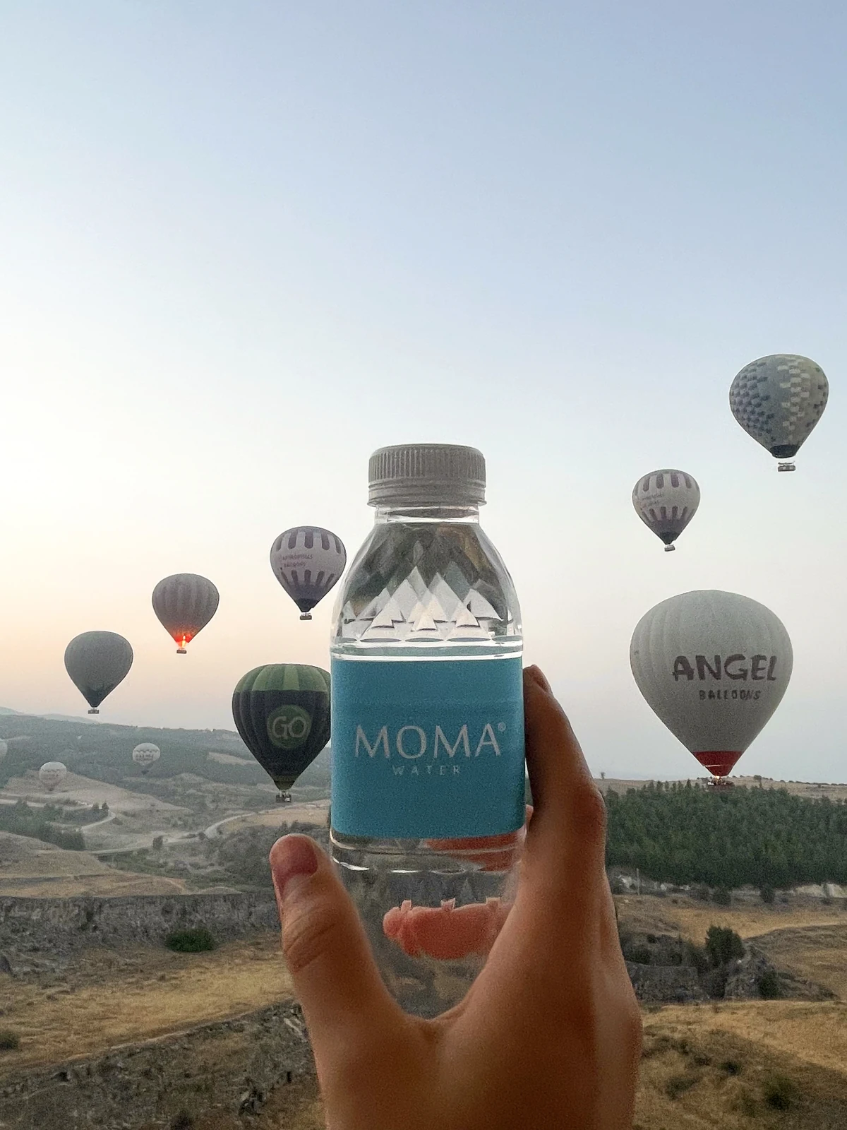 A MOMAwater bottle held to the sky as hot-air balloons rise at dawn.
